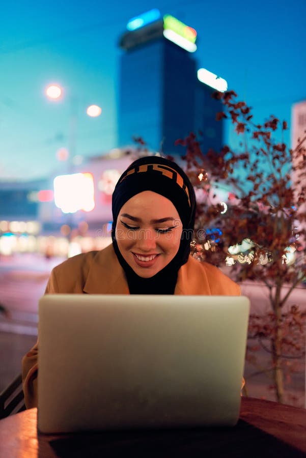 în atmosfera încântătoare a unui peisaj nocturn al orașului, o hijabclad-girl îngropată în laptop-ul ei crează o fascinantă stock images
