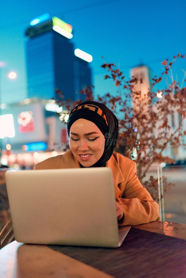 în atmosfera încântătoare a unui peisaj nocturn al orașului, o hijabclad-girl îngropată în laptop-ul ei crează o fascinantă stock images