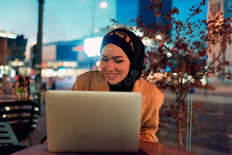 în atmosfera încântătoare a unui peisaj nocturn al orașului, o hijabclad-girl îngropată în laptop-ul ei crează o fascinantă stock photos