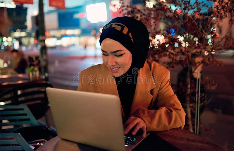 în atmosfera încântătoare a unui peisaj nocturn al orașului, o hijabclad-girl îngropată în laptop-ul ei crează o fascinantă royalty free stock images