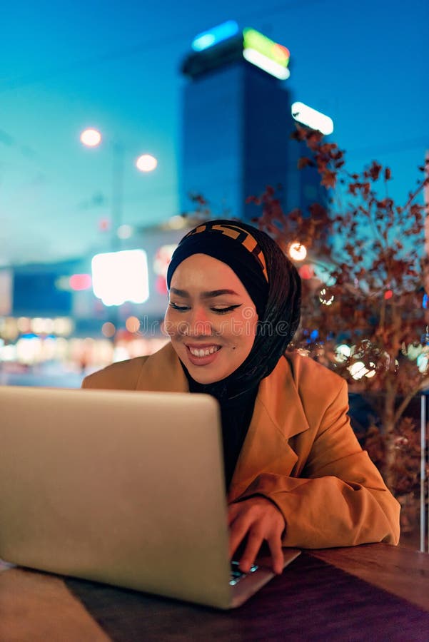 în atmosfera încântătoare a unui peisaj nocturn al orașului, o hijabclad-girl îngropată în laptop-ul ei crează o fascinantă royalty free stock image