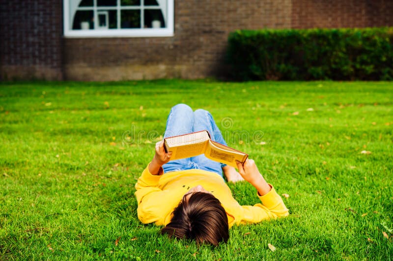 Enfant caucasien look décontracté en jean bleu et hoodie jaune pondre sur herbe verte profiter lire des histoires amusantes dans l image stock