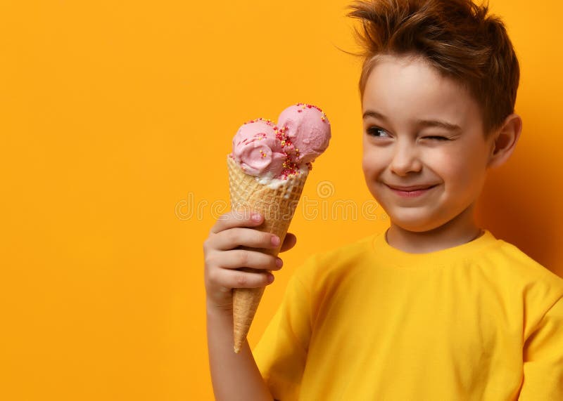Enfant de bébé garçon mangeant la crème glacée de fraise dans le cône de gaufres et clignant de l'oeil sur le jaune image stock Enfant de bébé garçon mangeant la crème glacée de fraise dans le cône de gaufres et clignant de l'oeil sur le jaune image stock