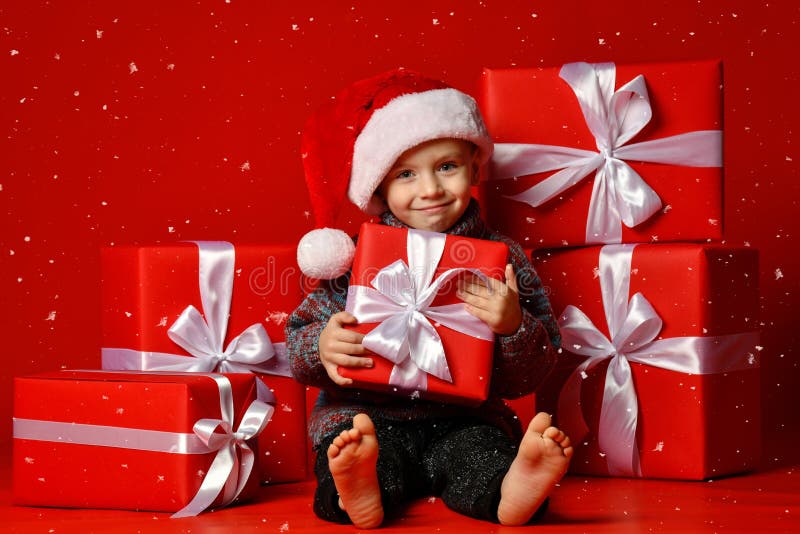 Enfant drôle de sourire dans le chapeau rouge de Santa jugeant le cadeau de Noël disponible Concept de Noël photo stock Enfant drôle de sourire dans le chapeau rouge de Santa jugeant le cadeau de Noël disponible Concept de Noël photo stock