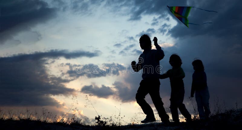Enfants jouant le cerf-volant sur le pré de coucher du soleil d'été silhouetté image stock Enfants jouant le cerf-volant sur le pré de coucher du soleil d'été silhouetté image stock
