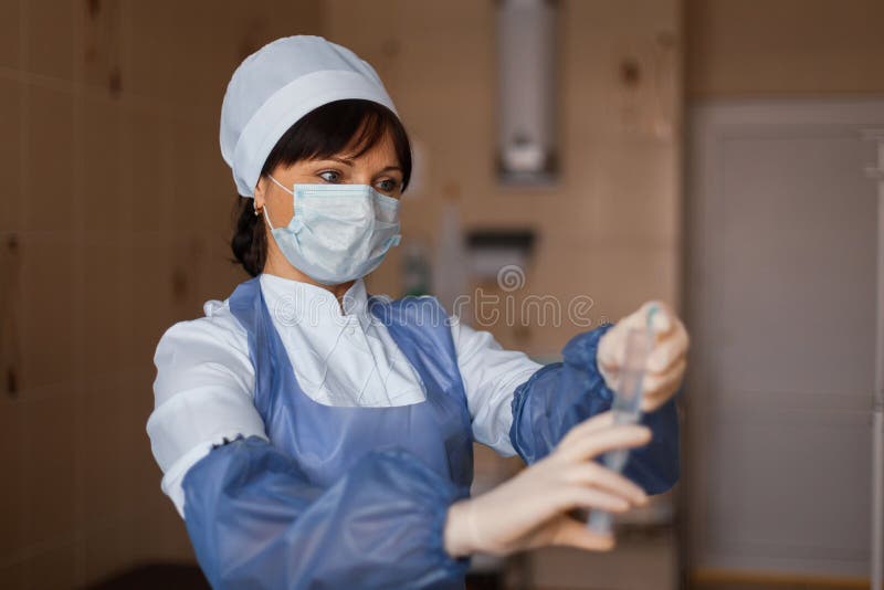Enfermeira em uma máscara branca nos suportes médicos azuis da roupa que guardam uma seringa em sua mão imagem de stock royalty free Enfermeira em uma máscara branca nos suportes médicos azuis da roupa que guardam uma seringa em sua mão imagem de stock royalty free