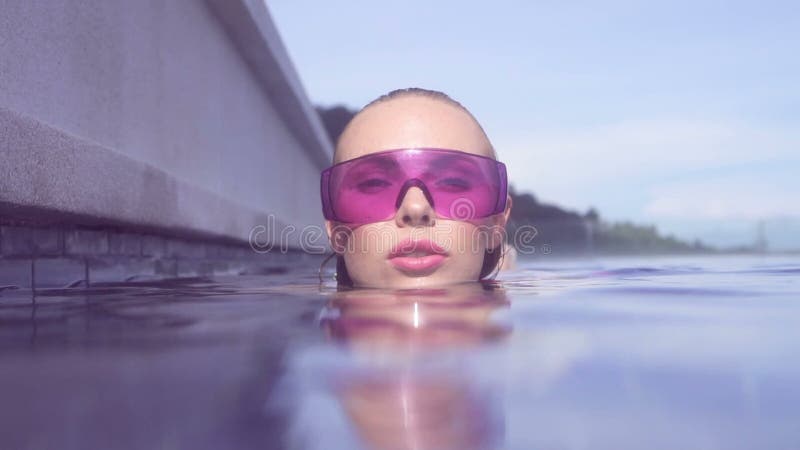 Enfrente o close up da jovem mulher 'sexy' que veste os óculos de sol roxos que olham a câmera na piscina do telhado da infinidad