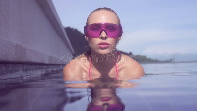 Enfrente o close up da jovem mulher 'sexy' que veste os óculos de sol roxos que olham a câmera na piscina do telhado da infinidad