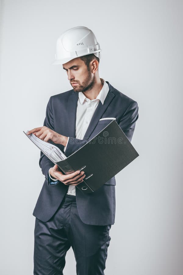 Handsome male in suit and hardhat holding big folder and flipping its pages while standing on light gray background. Handsome male in suit and hardhat holding big folder and flipping its pages while standing on light gray background