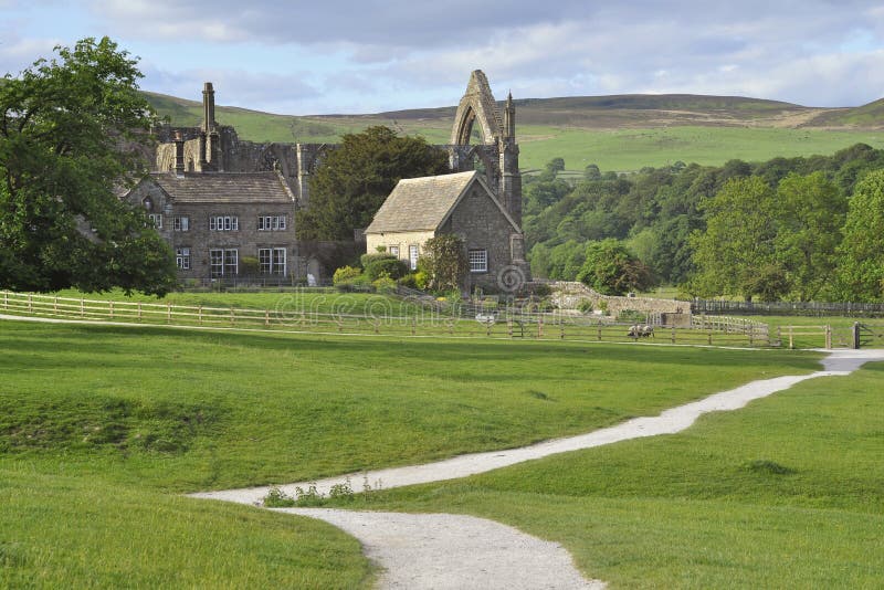 L'inglese in un paesaggio con un sentiero che conduce alle rovine dell'Abbazia di Bolton, Yorkshire Dales, circondato da edifici di fattoria, gli alberi, i boschi sulle colline e i campi erbosi, preso in una giornata di sole, con un cielo nuvoloso con schiarite.