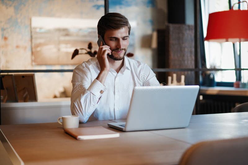 Confident young man working on laptop and talking on the mobile phone while sitting at his working place in office royalty free stock images Confident young man working on laptop and talking on the mobile phone while sitting at his working place in office royalty free stock images