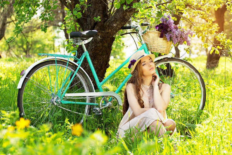 Enjoying nature. Horizontal shot of beautiful young woman keeping eyes closed while sitting on grass with retro bike near tree.