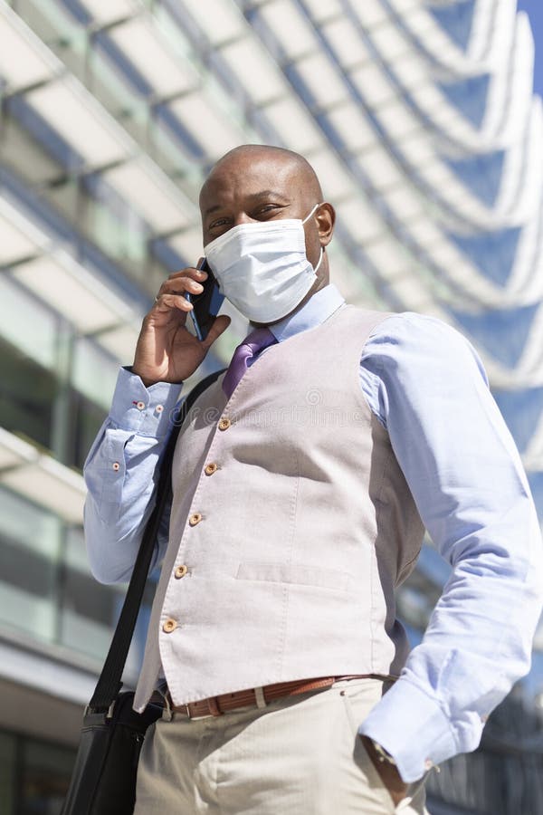 Enterprising black man in elegant clothes and medical mask is talking on the phone next to an office building