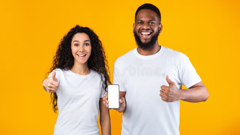 Excited Multiracial Couple Showing Phone Blank Screen Gesturing Thumbs Up In Approvement Standing Posing Over Yellow Studio Background. We Like This Mobile Application. Panorama, Mockup. Excited Multiracial Couple Showing Phone Blank Screen Gesturing Thumbs Up In Approvement Standing Posing Over Yellow Studio Background. We Like This Mobile Application. Panorama, Mockup