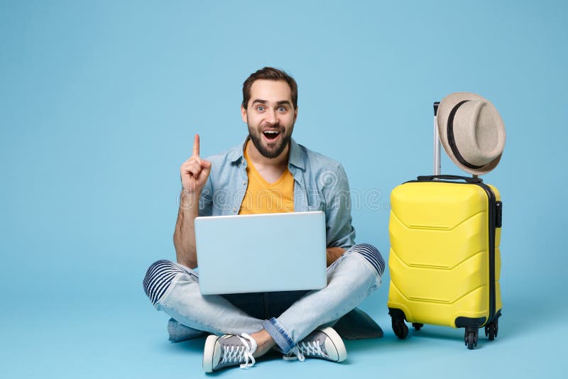 Excited traveler tourist man in yellow clothes isolated on blue background. Passenger traveling abroad on weekend. Air flight journey concept. Sit near suitcase, work on laptop, point index finger up. Excited traveler tourist man in yellow clothes isolated on blue background. Passenger traveling abroad on weekend. Air flight journey concept. Sit near suitcase, work on laptop, point index finger up