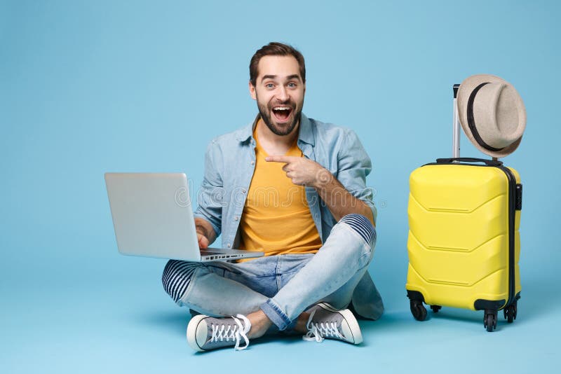 Excited traveler tourist man in yellow summer clothes isolated on blue background. Passenger traveling abroad on weekend. Air flight journey concept. Sit near suitcase pointing index finger on laptop. Excited traveler tourist man in yellow summer clothes isolated on blue background. Passenger traveling abroad on weekend. Air flight journey concept. Sit near suitcase pointing index finger on laptop