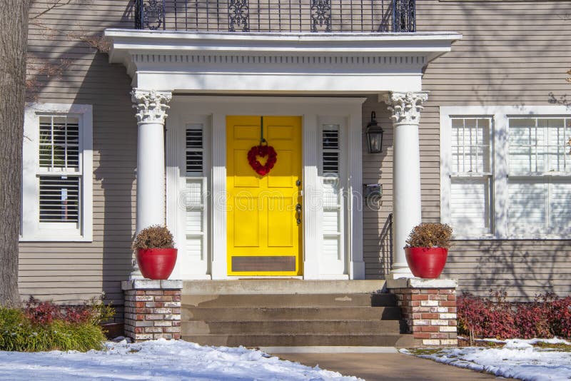 Entrance of pretty vintage house with ornate columns on porch and red valentine wreath on bright yellow door in snow