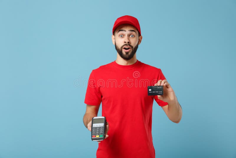 Delivery man in red workwear hold bank payment terminal to process acquire credit card payments isolated on blue background. Employee in cap t-shirt working courier. Service concept Mockup copy space. Delivery man in red workwear hold bank payment terminal to process acquire credit card payments isolated on blue background. Employee in cap t-shirt working courier. Service concept Mockup copy space