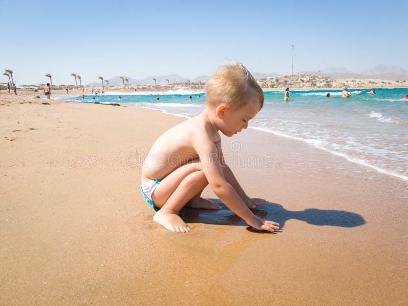Entz?ckende 3 Jahre alte Kleinkindjunge, die auf dem Seestrand in den warmen ruhigen Wellen sitzen und mit nass Sand spielen Ents stockbild Entz?ckende 3 Jahre alte Kleinkindjunge, die auf dem Seestrand in den warmen ruhigen Wellen sitzen und mit nass Sand spielen Ents stockbild