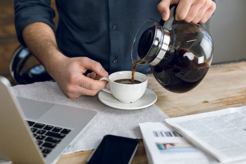 Equipaggi il caffè di versamento in tazza sullo scrittorio con il computer portatile e lo smartphone