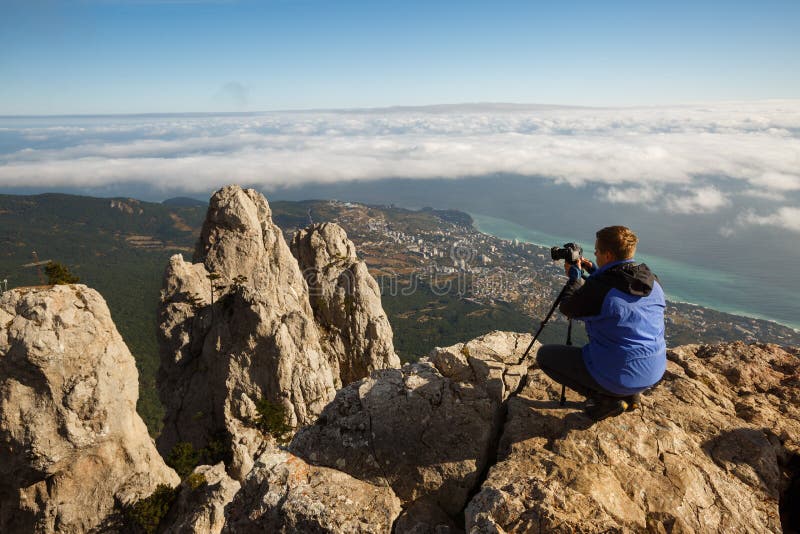 Equipaggi la seduta con una macchina fotografica della foto e del treppiede su un picco di alta montagna sopra le nuvole, la citt fotografie stock Equipaggi la seduta con una macchina fotografica della foto e del treppiede su un picco di alta montagna sopra le nuvole, la citt fotografie stock
