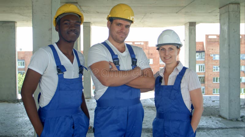 Equipo diverso confiado de trabajadores y de mujeres que se colocan agrupados en sus overoles y cascos de protección que sonríe e fotografía de archivo Equipo diverso confiado de trabajadores y de mujeres que se colocan agrupados en sus overoles y cascos de protección que sonríe e fotografía de archivo