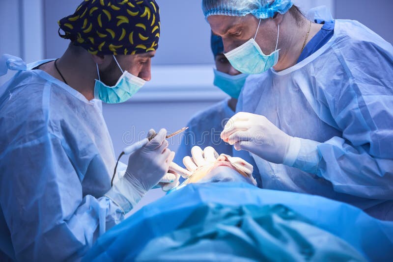 Equipo quirúrgico que realiza cirugía en el teatro de operación moderno, equipo de doctores que concentran en un paciente durante imagen de archivo Equipo quirúrgico que realiza cirugía en el teatro de operación moderno, equipo de doctores que concentran en un paciente durante imagen de archivo