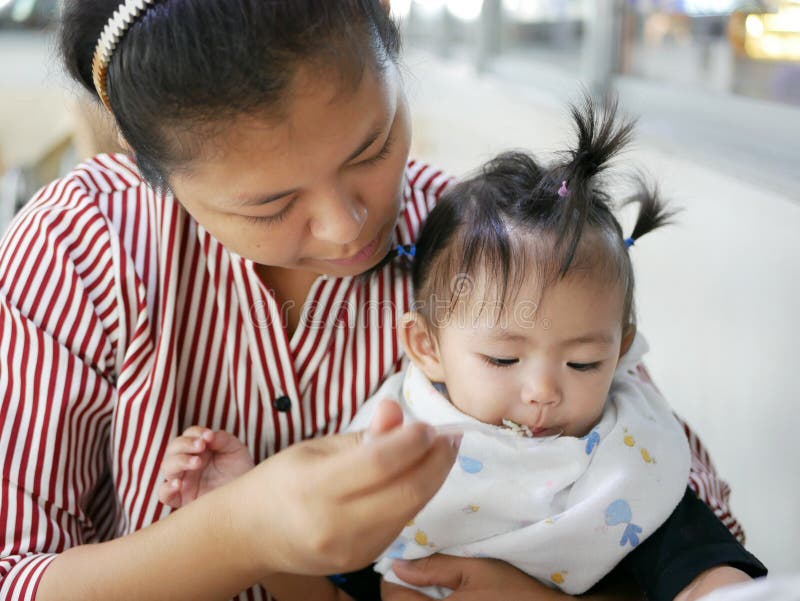 Erfahrene asiatische Mutter, die kleinen Löffel verwendet, um ihr Baby, 12 Monate alte, an einer Cafeteria einzuziehen stockbild Erfahrene asiatische Mutter, die kleinen Löffel verwendet, um ihr Baby, 12 Monate alte, an einer Cafeteria einzuziehen stockbild