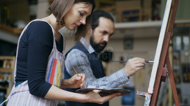 Erfahrener Künstlermann, der Malerei der jungen Frau auf Gestell am Kunstakademiestudio - Kreativitäts-, Bildungs- und Kunstleute stockfoto Erfahrener Künstlermann, der Malerei der jungen Frau auf Gestell am Kunstakademiestudio - Kreativitäts-, Bildungs- und Kunstleute stockfoto