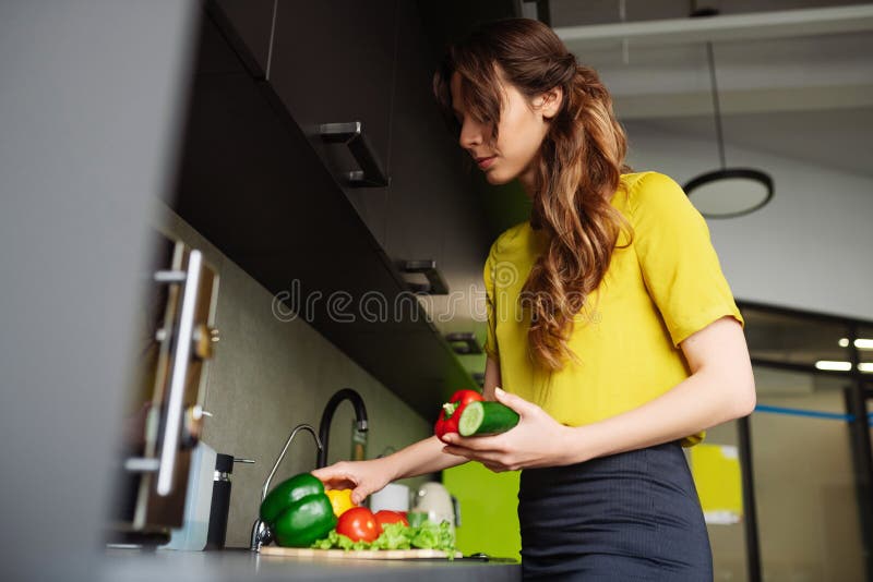 Vitamin food. Satisfied woman in short-sleeve blouse standing in the kitchen with dark furniture picking up washed vegetables in her hands. Vitamin food. Satisfied woman in short-sleeve blouse standing in the kitchen with dark furniture picking up washed vegetables in her hands