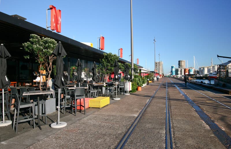 Modification of repository restored into sightseeing destination in North Wharf, Wynyard Quarter, Auckland, New Zealand. Redevelopment of cargo storehouse converted into restaurants with outdoor seating, and abandoned train rails on pedestrian boardwalk. Transformation of old building redeveloped into eateries with exterior seats and tables, and decommissioned rail tracks on promenade. Remodeling of dockside depository transformed into cafes with outside chairs and stools, and railroad tracks on pathway. Conversion of freight depot rebuilt into popular tourist attraction in docklands. Restoration of storage architecture renovated into bars. Rebuilding of architectural landmark modified into dinning. road street path walk passage passageway. Modification of repository restored into sightseeing destination in North Wharf, Wynyard Quarter, Auckland, New Zealand. Redevelopment of cargo storehouse converted into restaurants with outdoor seating, and abandoned train rails on pedestrian boardwalk. Transformation of old building redeveloped into eateries with exterior seats and tables, and decommissioned rail tracks on promenade. Remodeling of dockside depository transformed into cafes with outside chairs and stools, and railroad tracks on pathway. Conversion of freight depot rebuilt into popular tourist attraction in docklands. Restoration of storage architecture renovated into bars. Rebuilding of architectural landmark modified into dinning. road street path walk passage passageway