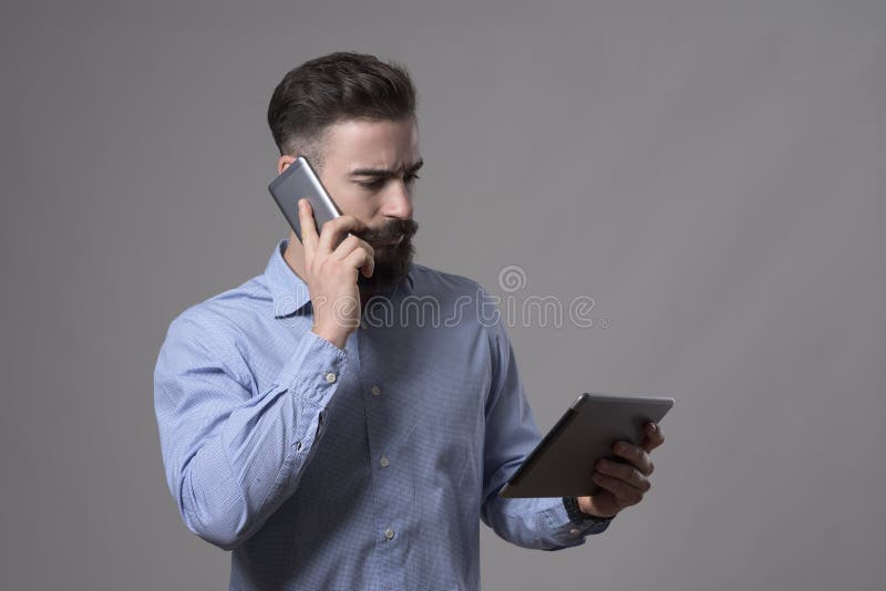 Ernster überzeugter junger Geschäftsmann, der am Telefon spricht und Tablet-Computer betrachtet lizenzfreie stockfotografie Ernster überzeugter junger Geschäftsmann, der am Telefon spricht und Tablet-Computer betrachtet lizenzfreie stockfotografie