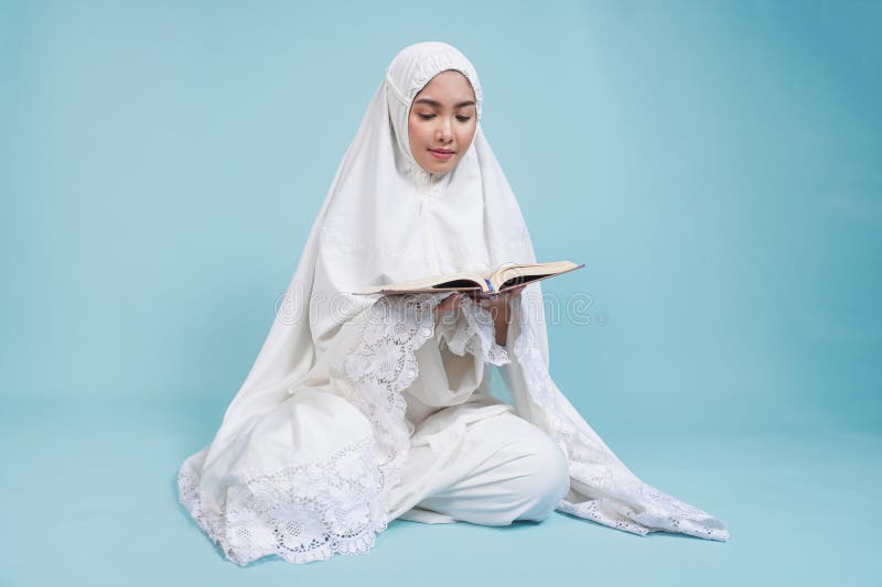 Ernstzunehmende junge asiatische Muslimfrau im Gebetskleid sitzt herunter und liest Quran über isolierten blauen Hintergrund. Hads lizenzfreie stockbilder