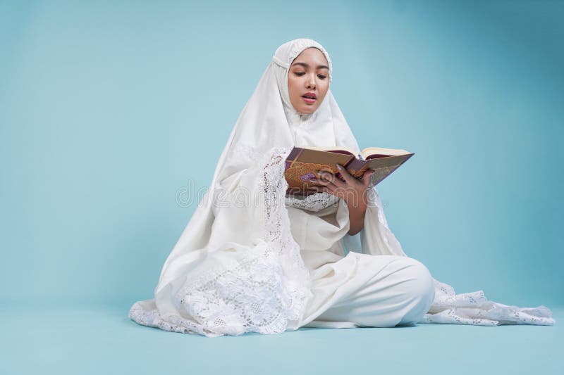 Ernstzunehmende junge asiatische Muslimfrau im Gebetskleid sitzt herunter und liest Quran über isolierten blauen Hintergrund. Hads stockfotos