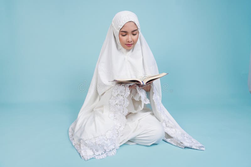 Ernstzunehmende junge asiatische Muslimfrau im Gebetskleid sitzt herunter und liest Quran über isolierten blauen Hintergrund. Hads stockfotografie