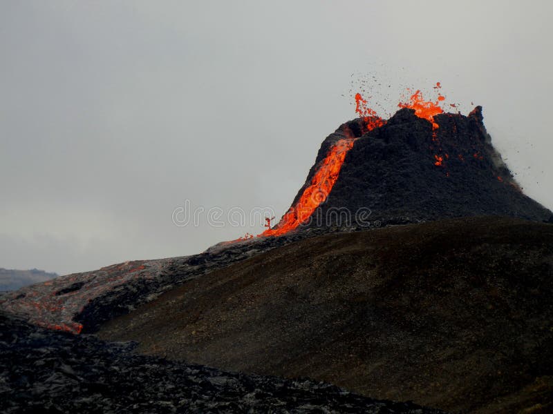 Active Geldingadalir volcano in Iceland is erupting with a stream of lava flowing out of the crater. The hot orange and grey lava forms a river flowing through older, cold black lava. Barren land, black lava, grey volcano and gloomy skies contrasting with the bring orange lava stream. Photo taken 23.3.2021. Active Geldingadalir volcano in Iceland is erupting with a stream of lava flowing out of the crater. The hot orange and grey lava forms a river flowing through older, cold black lava. Barren land, black lava, grey volcano and gloomy skies contrasting with the bring orange lava stream. Photo taken 23.3.2021.