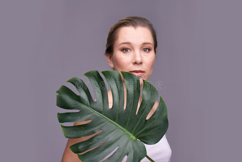 Erwachsene kaukasische Weibchen, die mit Monstera-Blatt vor grauem Hintergrund posieren stockfoto