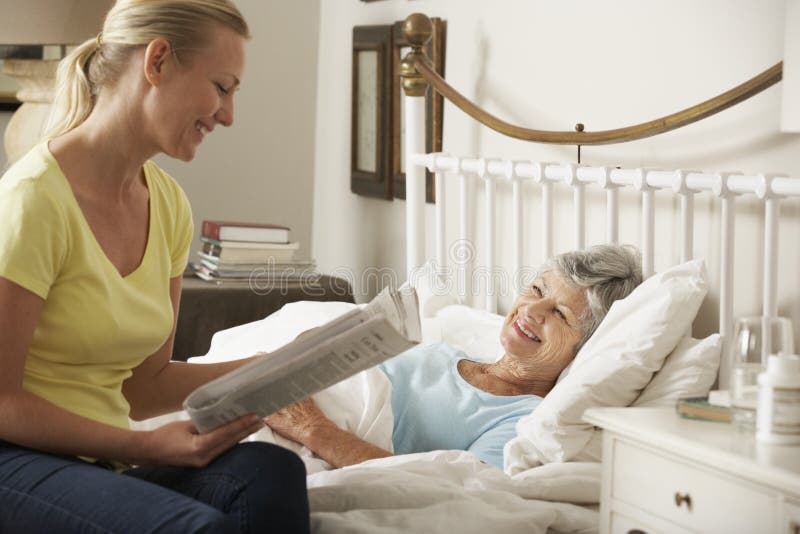 Erwachsene Tochter-Lesezeitung zum älteren weiblichen Elternteil im Bett zu Hause stockfotos Erwachsene Tochter-Lesezeitung zum älteren weiblichen Elternteil im Bett zu Hause stockfotos