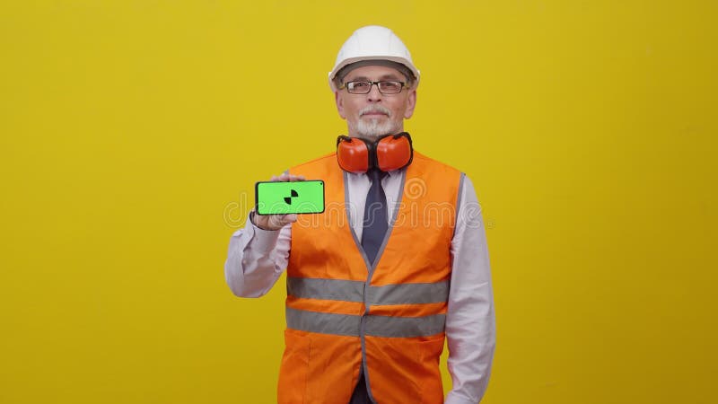 Erwachsener Mann Ingenieur in Arbeitsuniform hält Handy mit App und schaut in die Kamera lächelnd gegen die gelbe