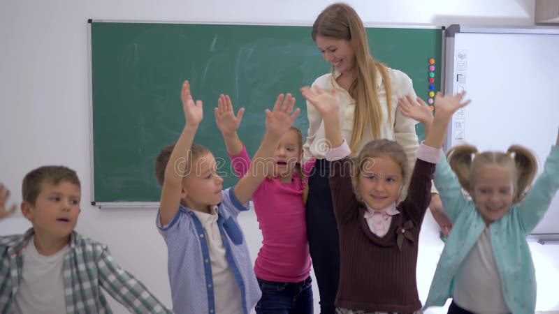 Escola primária, grupo de mãos de salto e de ondulação do divertimento das crianças próximo ao professor no fundo da placa