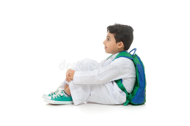 Escolar árabe que se sienta en la tierra con una sonrisa en su cara, saudí tradicional blanco que lleva Thobe, mochila y zapatill