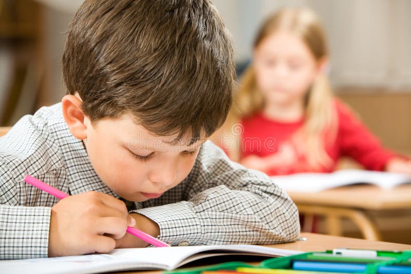 Photo of clever schoolkid writing something in his copybook at lesson on background of classmate. Photo of clever schoolkid writing something in his copybook at lesson on background of classmate