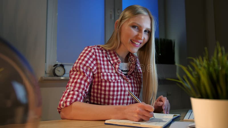 Escritura femenina alegre en planificador diario Mujer rubia joven animada en la camisa a cuadros casual que se sienta en el escr