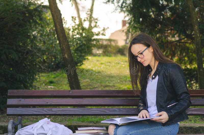 Escuela o estudiante universitaria hermosa joven con los vidrios que se sientan en el banco en el parque que lee los libros y el fotos de archivo libres de regalías Escuela o estudiante universitaria hermosa joven con los vidrios que se sientan en el banco en el parque que lee los libros y el fotos de archivo libres de regalías