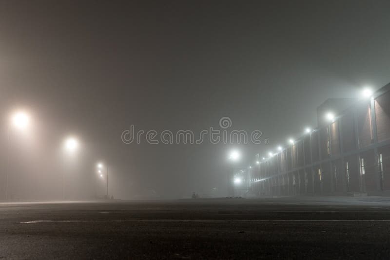Estacionamento urbano vazio e revérbero do carro na noite nevoenta Construção e lanternas industriais velhas de tijolo na rua só fotografia de stock royalty free Estacionamento urbano vazio e revérbero do carro na noite nevoenta Construção e lanternas industriais velhas de tijolo na rua só fotografia de stock royalty free
