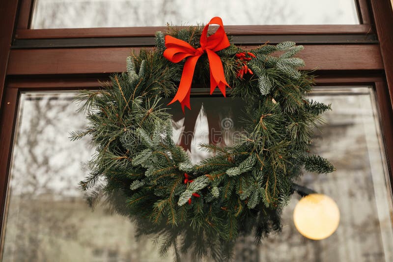 Estiloso corona de Navidad con arco rojo y bayas delante de la tienda en el mercado de vacaciones en la calle de la ciudad. Espaci imagen de archivo libre de regalías Estiloso corona de Navidad con arco rojo y bayas delante de la tienda en el mercado de vacaciones en la calle de la ciudad. Espaci imagen de archivo libre de regalías