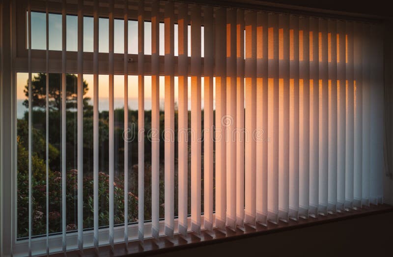 Estores brancas verticais da barra pendurados em frente a uma janela enquanto o sol se põe a tornar a luz dourada