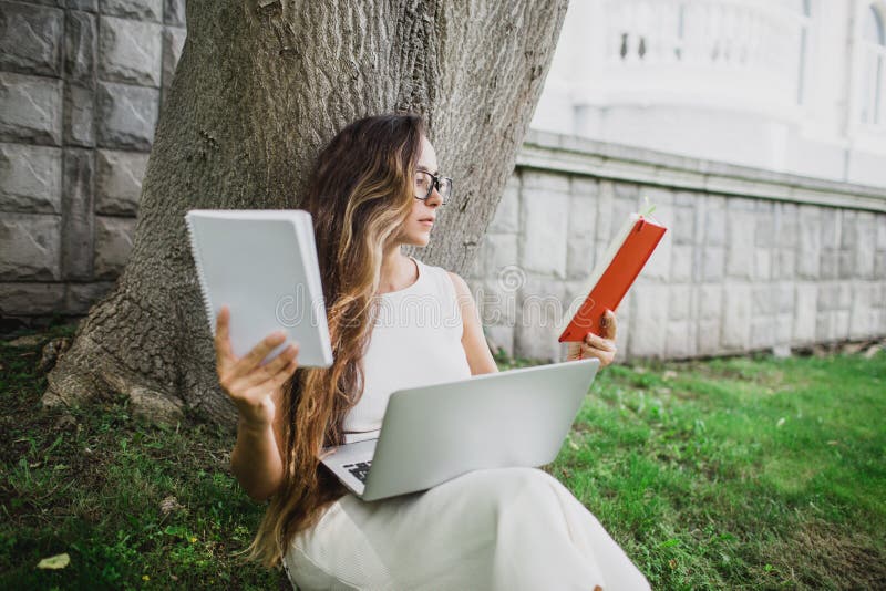 Beautiful girl student learning her lessons using her laptop and notes, sitting on green lawn in a yard of university. Beautiful girl student learning her lessons using her laptop and notes, sitting on green lawn in a yard of university.