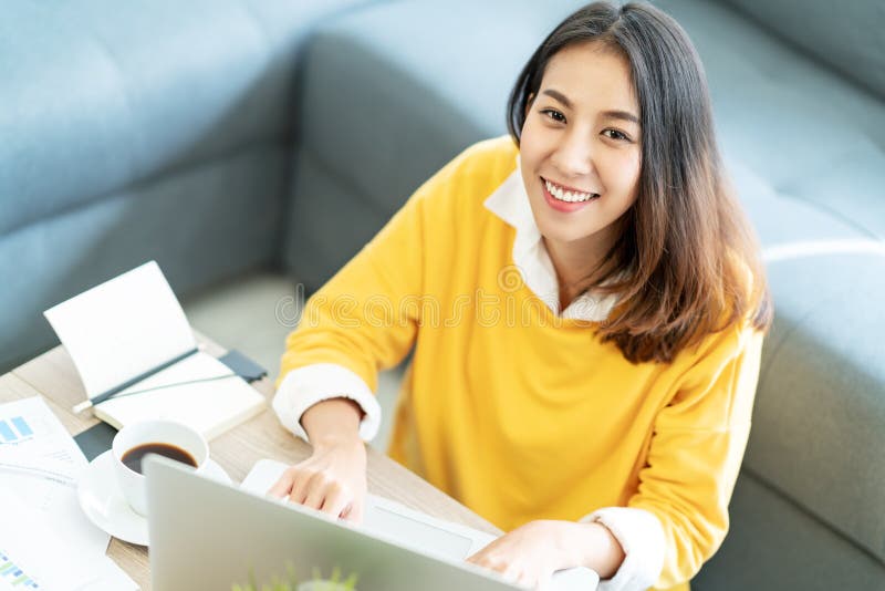 Estudante fêmea asiático feliz atrativo novo que senta-se no assoalho da sala de visitas que sorri e que olha acima na câmera imagem de stock royalty free Estudante fêmea asiático feliz atrativo novo que senta-se no assoalho da sala de visitas que sorri e que olha acima na câmera imagem de stock royalty free