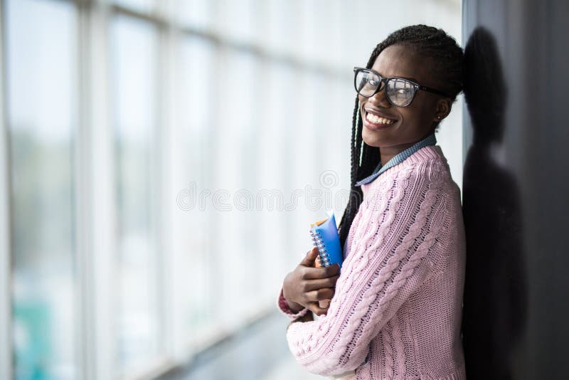 Estudiante afroamericana de la belleza joven en los vidrios que sostienen los cuadernos en pasillo moderno de la universidad imágenes de archivo libres de regalías Estudiante afroamericana de la belleza joven en los vidrios que sostienen los cuadernos en pasillo moderno de la universidad imágenes de archivo libres de regalías